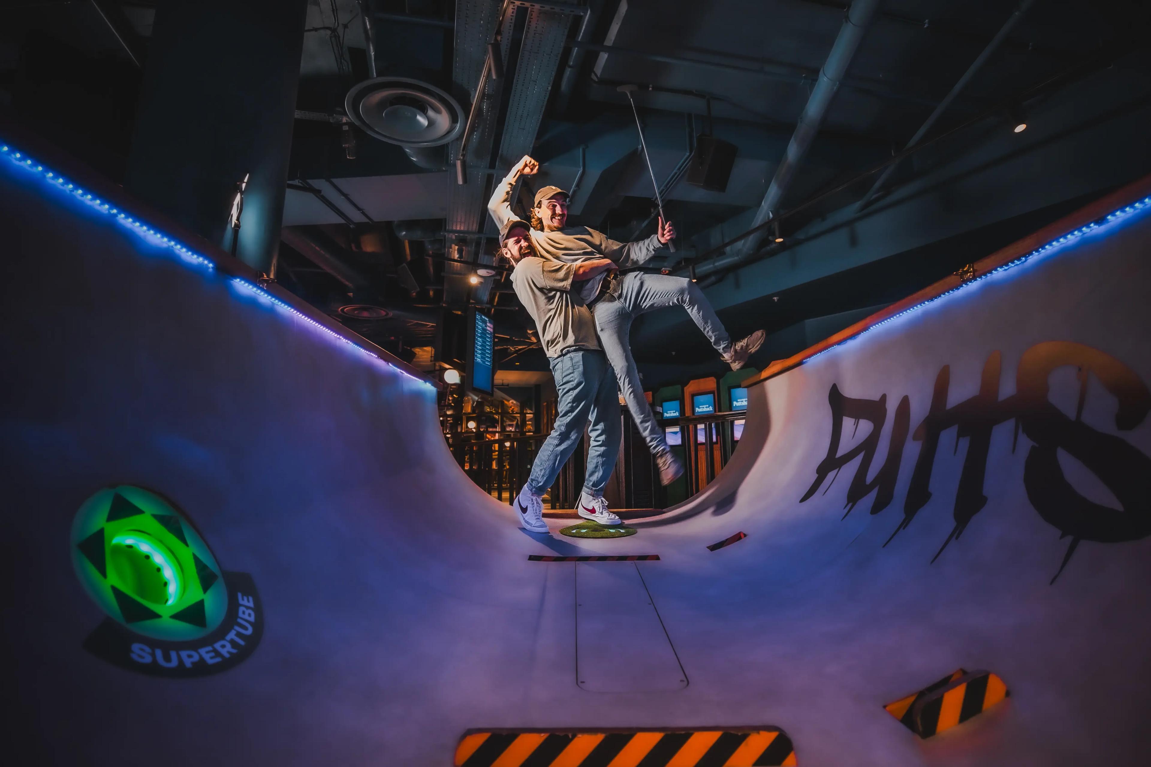Two friends celebrating on the half-pipe skateboarding ramp hole at a Puttshack
