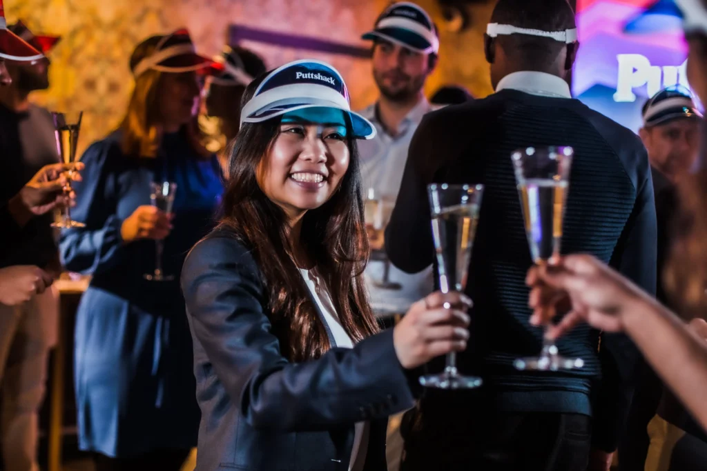 A smiling woman wearing a Puttshack blue visor hold a fancy glass