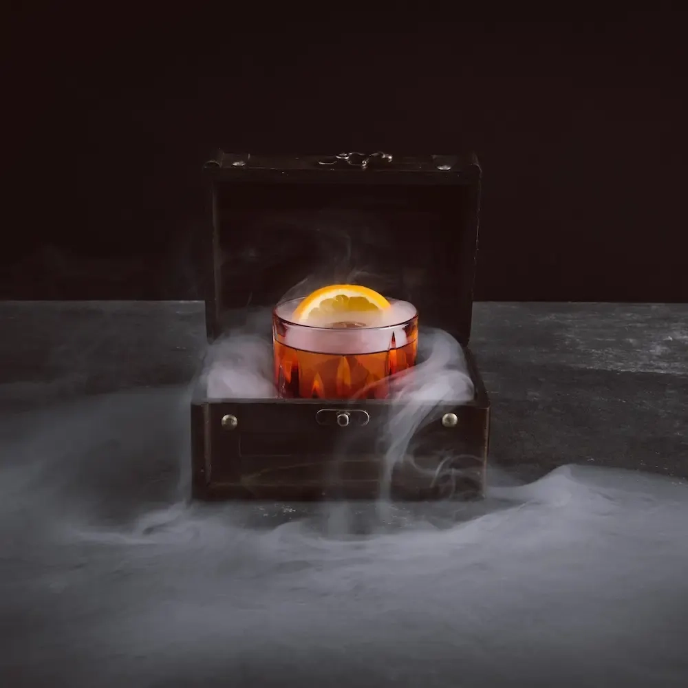 A dark alcoholic drink with a citrus slice sitting in a box that is smoking