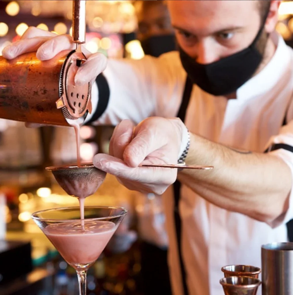 A bartender that's wearing a mask straining a cocktail into a glass