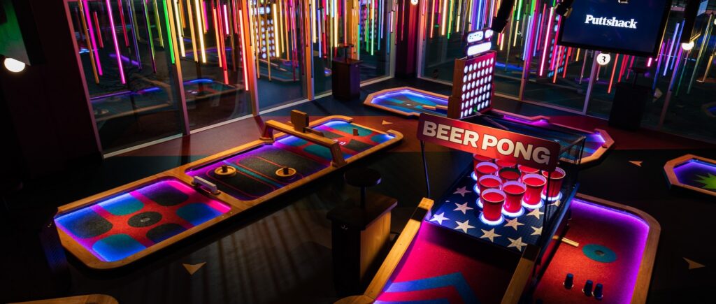 An indoor mini golf course featuring brightly lit putting lanes and a beer pong–style obstacle with red cups, surrounded by colorful neon lights and illuminated course elements