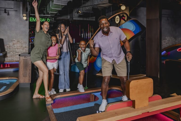 A group of adults and children standing on an indoor mini golf course, holding mini golf clubs and gathered around a putting lane with colorful obstacles and mural artwork visible in the background