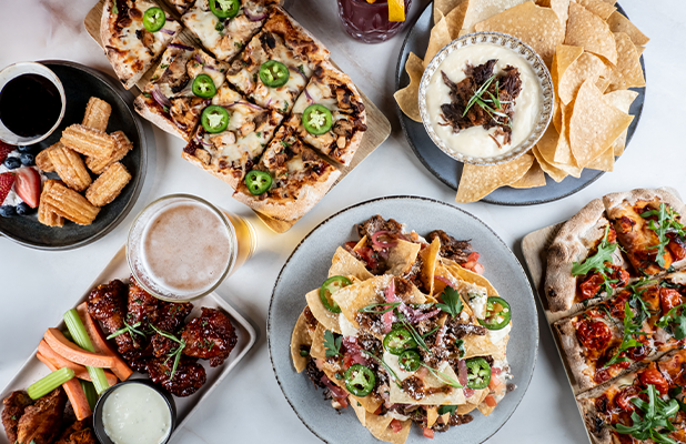 Overhead view of assorted shareable dishes including flatbread pizza, nachos, chicken wings with vegetables and dipping sauce, tortilla chips with dip, and drinks on a table