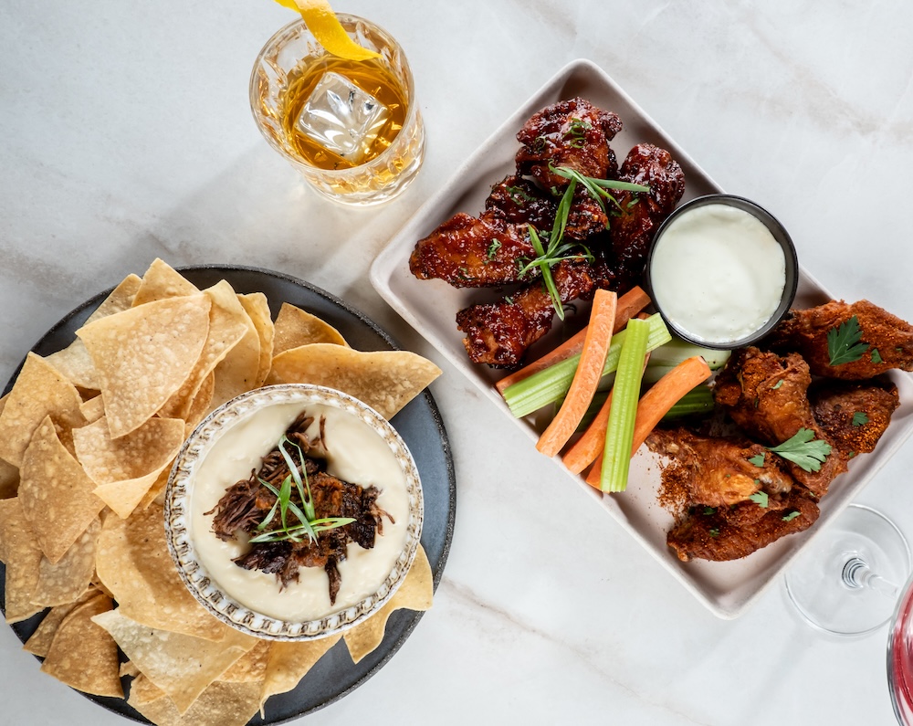Overhead view of shareable appetizers including chicken wings with celery and carrot sticks, tortilla chips with a dipped appetizer, fried bites, and drinks arranged on a table