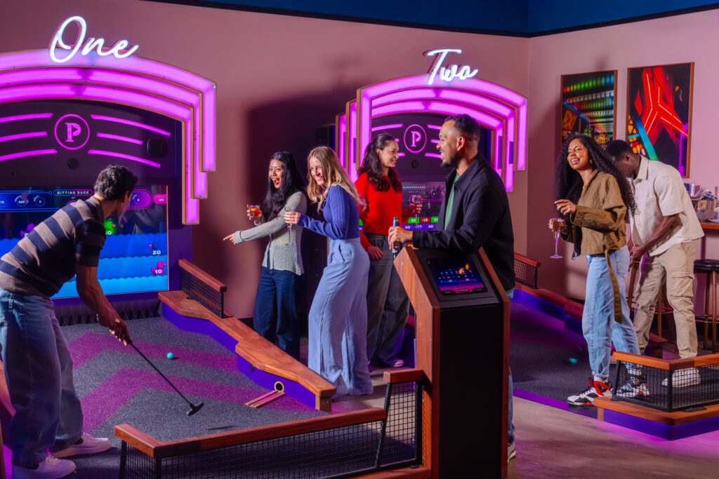 A group of young adults laughing and joking while playing on both Puttshack Puttcade bays