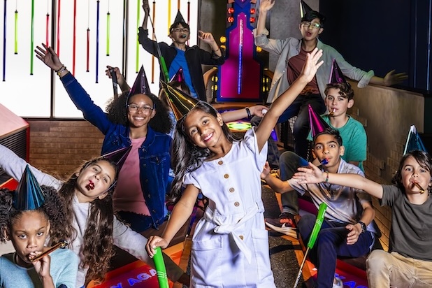 A large group on an indoor mini golf course wearing party hats and holding noisemakers, with several people raising their arms and mini golf clubs under colorful lighting