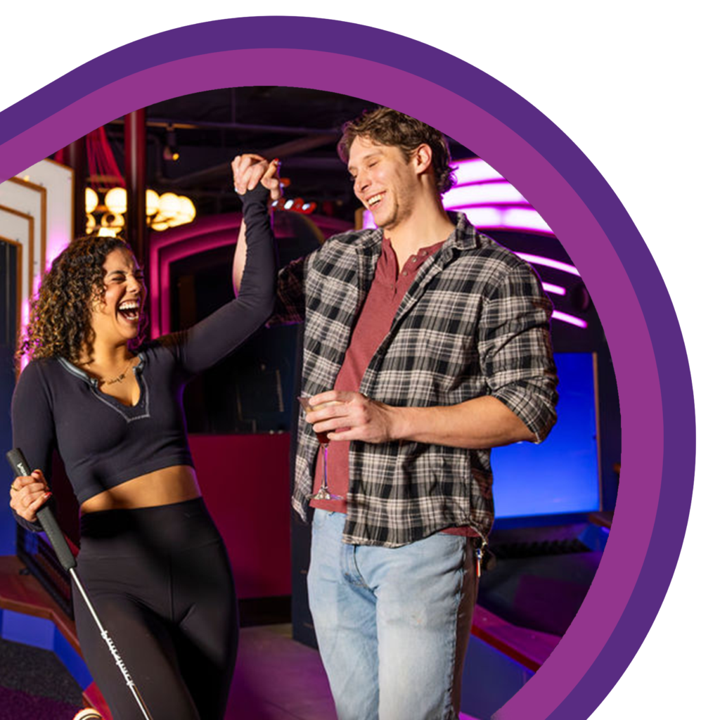 Two people standing on an indoor mini golf course holding mini golf clubs and drinks, with one person raising the other’s hand beside a putting lane under colorful lighting
