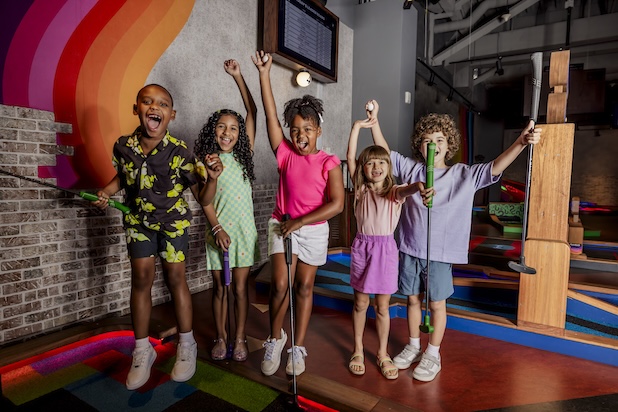 A group of children standing on an indoor mini golf course holding mini golf clubs, with several raising their arms and one holding a golf ball, set against a colorful mural and game elements in the background