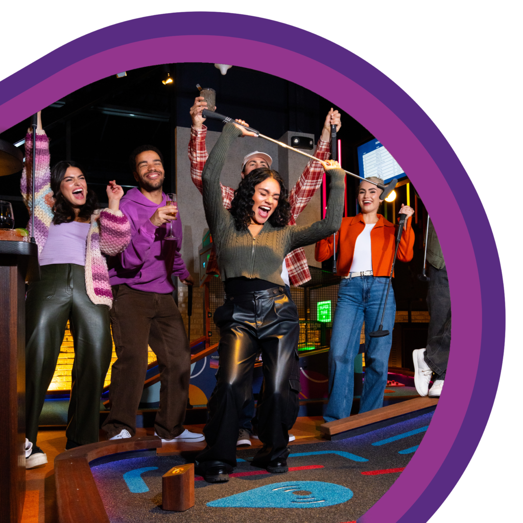 A group of people standing on an indoor mini golf course with raised mini golf clubs and drinks, gathered around a putting lane under colorful interior lighting