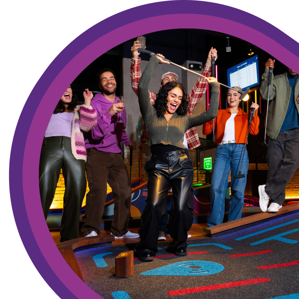 Group of people standing together on an indoor mini golf course with raised arms and mini golf putters during a group activity