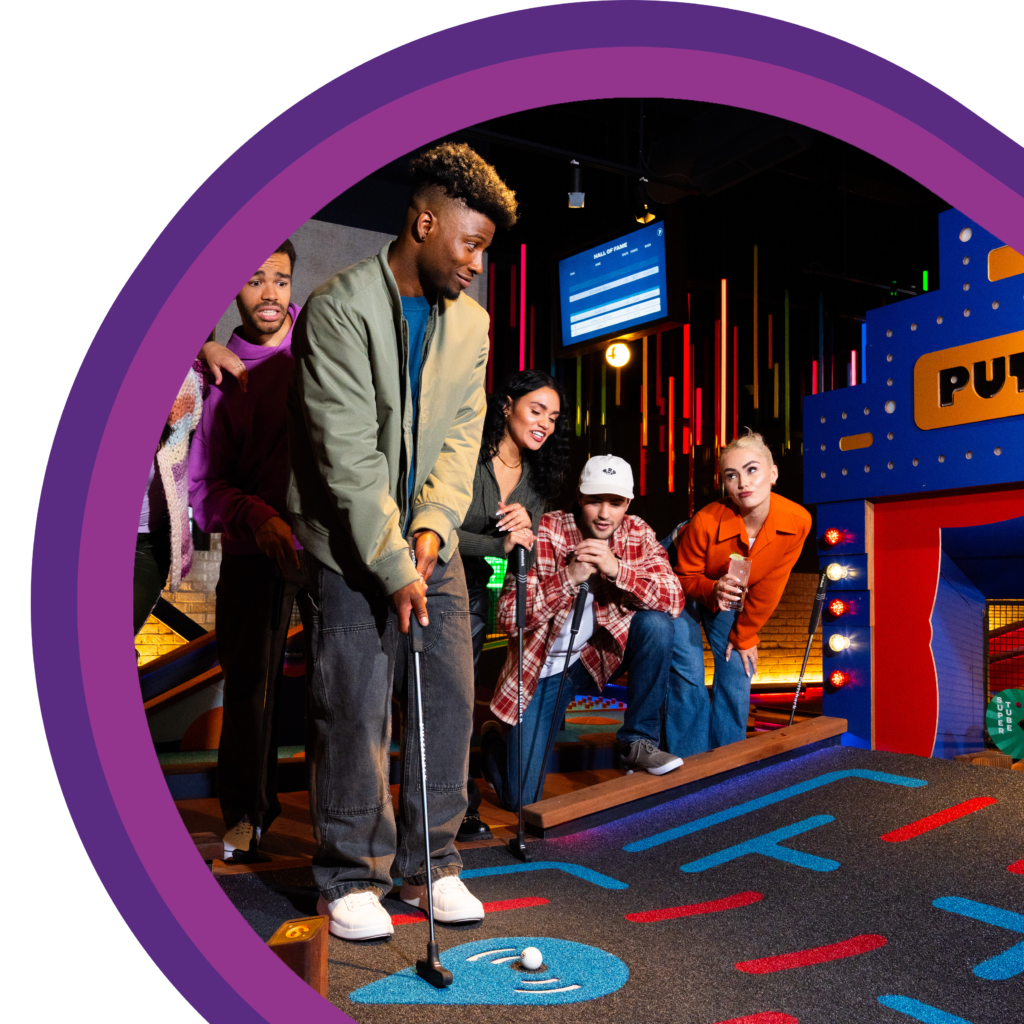 A group of people gathered around an indoor mini golf putting lane, with one person lining up a putt while others watch closely under colorful interior lighting