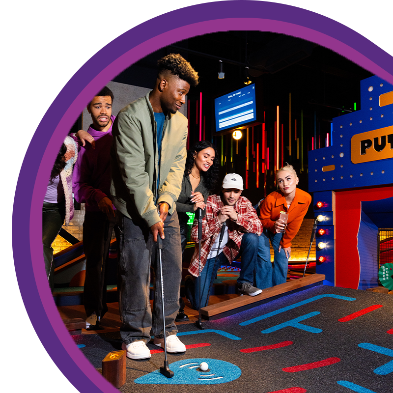 Group playing an indoor mini golf game with one person putting while others watch nearby
