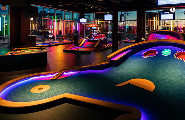 Wide view of an indoor mini golf venue featuring curved courses with illuminated edges and colorful lighting