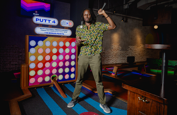 Person standing on an indoor mini golf course holding a putter next to a PUTT 4 game board with illuminated targets