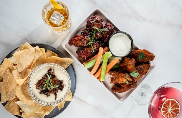 Assorted shareable appetizers on a table including chicken wings with vegetables and dipping sauce, tortilla chips with dip, and mixed cocktails