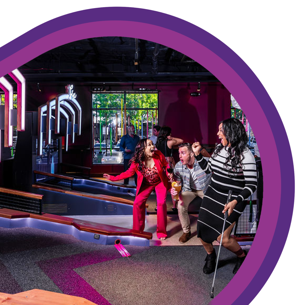 A group of people standing beside an indoor mini golf lane, holding mini golf clubs and gesturing toward the putting area under colorful interior lighting