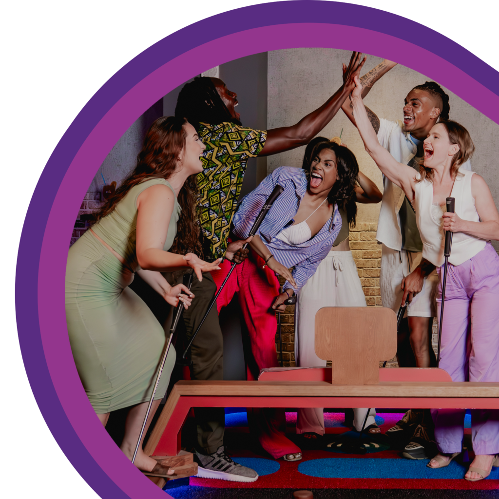 A group of people standing around an indoor mini golf obstacle and raising their hands for high-fives, with mini golf clubs visible and colorful lighting framing the scene
