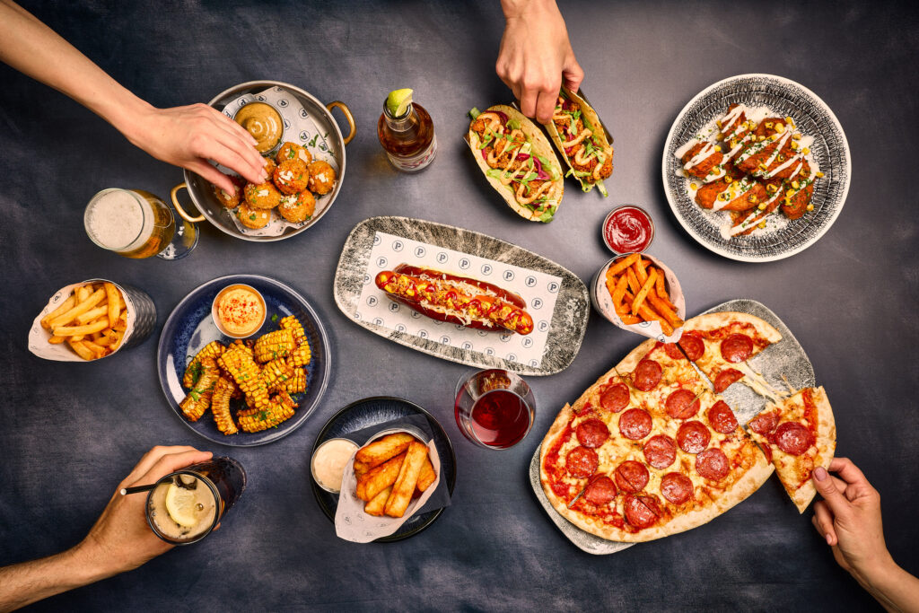 Hands reaching out to. try different food dishes like pizzas, tacos, hotdogs, fries